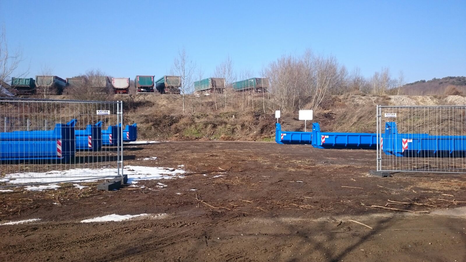 Blaue Abfallcontainer stehen auf einem mit einem Bauzaun abgesperrtem Platz. Im hinteren Bereich des Platzes befindet sich ein kleiner Hang, welcher etwas bewachsen ist. Auf der darüberliegenden Ebene stehen Lastkraftwagen, welche jeweils mit einem Abfallcontainer beladen sind.