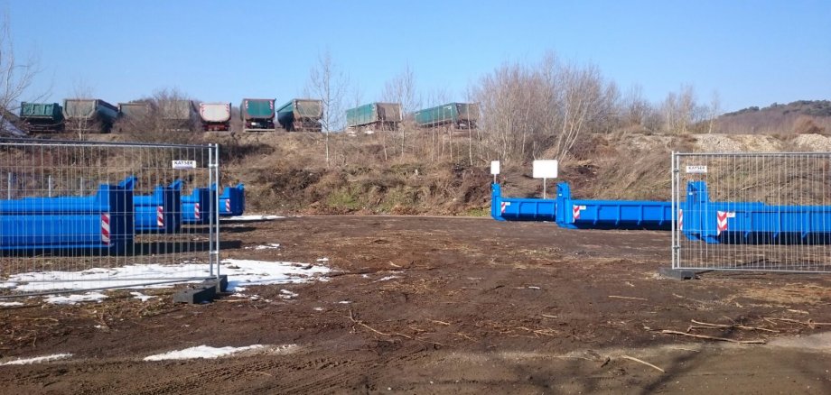 Blaue Abfallcontainer stehen auf einem mit einem Bauzaun abgesperrtem Platz. Im hinteren Bereich des Platzes befindet sich ein kleiner Hang, welcher etwas bewachsen ist. Auf der darüberliegenden Ebene stehen Lastkraftwagen, welche jeweils mit einem Abfallcontainer beladen sind.