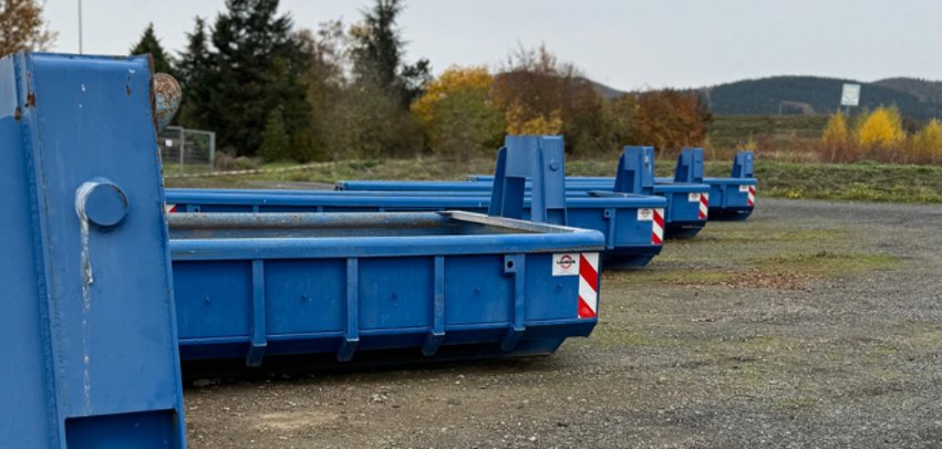 Blaue Abfallcontainer stehen auf einem mit einem Bauzaun abgesperrtem Platz.