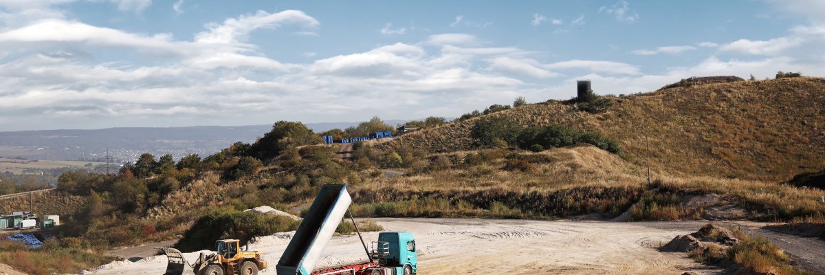 Sicht auf die Deponie Blick auf das weitläufige Deponiegelände. In der Mitte steht ein LKW, der Abfälle abkippt. Es ist blauer Himmel zu erkennen.