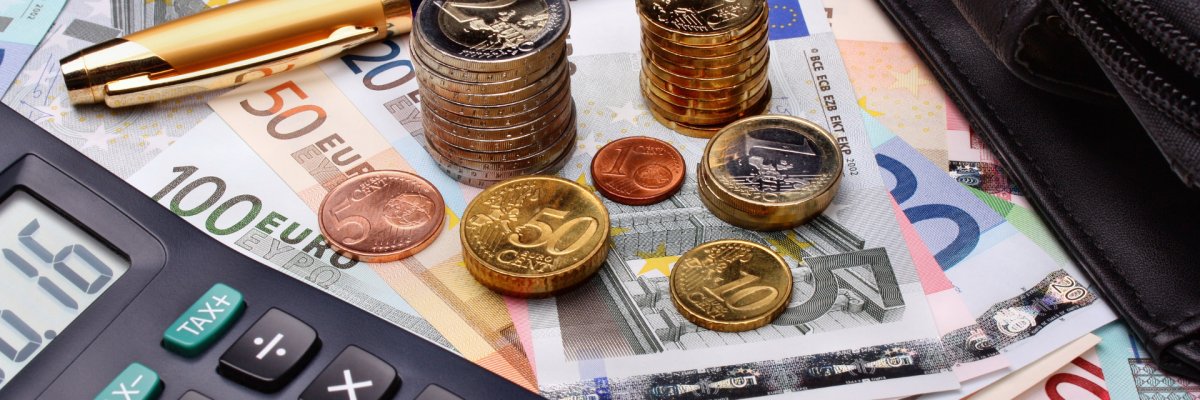 Mehrere Geldscheine und Münzen liegen auf einem Tisch. Daneben liegt ein Taschenrechner und ein Kugelschreiber. 