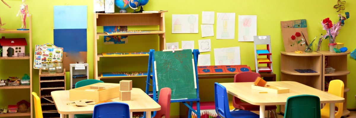 Blick in einen Gruppenraum eines Kindergartens mit grüner Wand im Hintergrund. Im Vordergrund stehen zwei Tische und mehrere Stühle. Auf den Tischen und drum herum liegen Spielzeuge. 