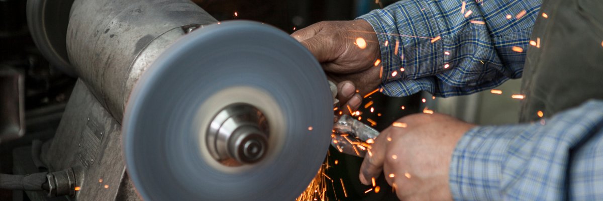 Handwerksbetrieb Ein Handwerker, dessen Hände zu sehen sind, hält ein Metallstück an ein sich drehendes Schleifgerät. Dadurch entsteht Funkenflug.