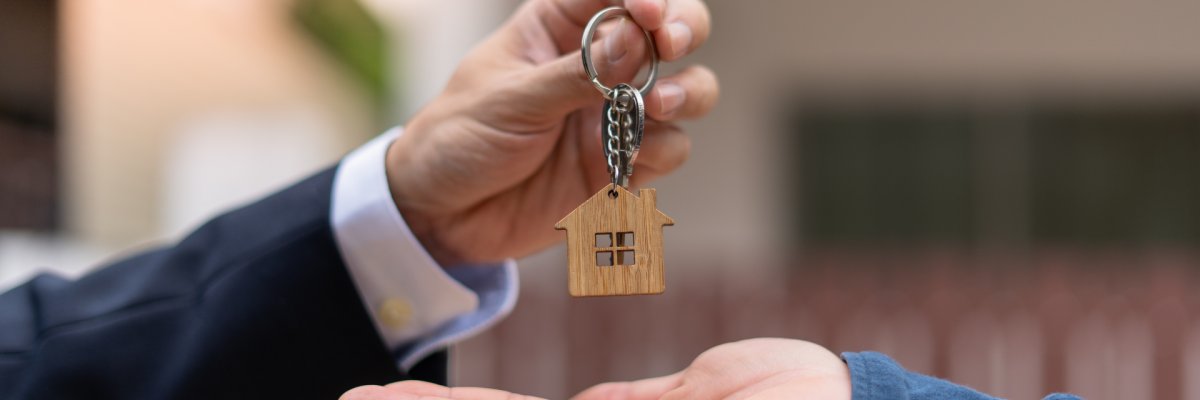 zwei in Sakko und Hemd gekleidete Arme und Hände, welche sich einen Schlüssel mit einem Schlüsselanhänger im Hausformat übergeben.  Die Handfläche der unteren Hand ist nach oben flach geöffnet, währenddessen die andere Hand den Schlüssel dort hinein legt. 