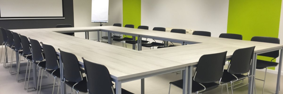 Blick in einen modern und grüner Wandbemalung gestalteten Sitzungssaal, in welchem mehrere Tische rechteckig zusammenstehen. Um die Tische stehen viele Stühle. Im Hintergrund ist eine Leinwand und eine Flipchart. 