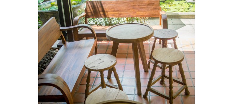 Gut erhaltene Möbel wie Barhocker, Tisch und Stühle stehen kreisförmig angeordnet auf mit Platten befestigtem Untergrund. Im Hintergrund ist ein Garten zu erkennen. 