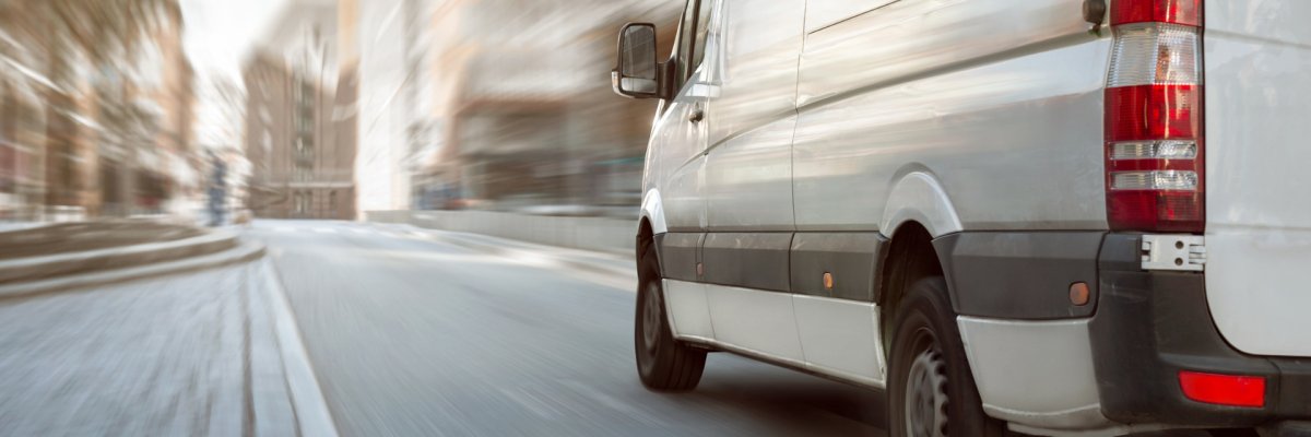 Ein weißer Transporter fährt auf einer asphaltierten Straße innerhalb der Stadt. Die Wohnhäuser sind verschwommen zu erkennen. 