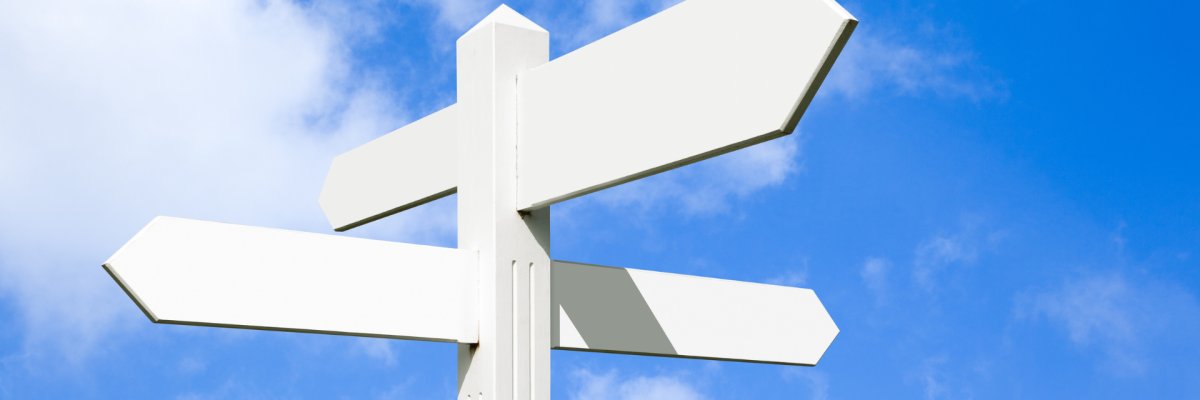 Ein großer Wegweiser, befindet sich mittig im Bild. Vier weiße Pfeile zeigen in verschiedene Richtungen. Die Pfeile sind nicht beschriftet. Im Hintergrund ist blauer Himmel mit ein paar dünnen Wölkchen zu sehen.